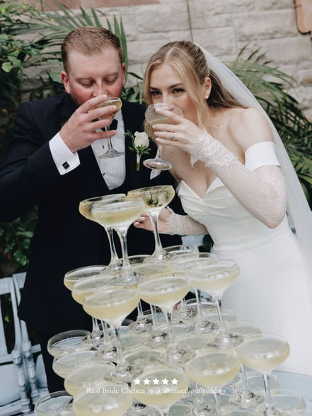 Wedding couple toasting with a pyramid of champagne glasses. Bride wearing a Harriett Falvey veil and double edged lace bridal gloves.