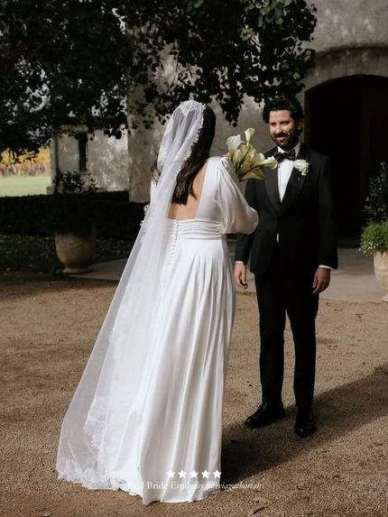 Woman in a white wedding dress and chantilly lace mantilla veil standing next to a man in a black suit with a bow tie, outdoors.